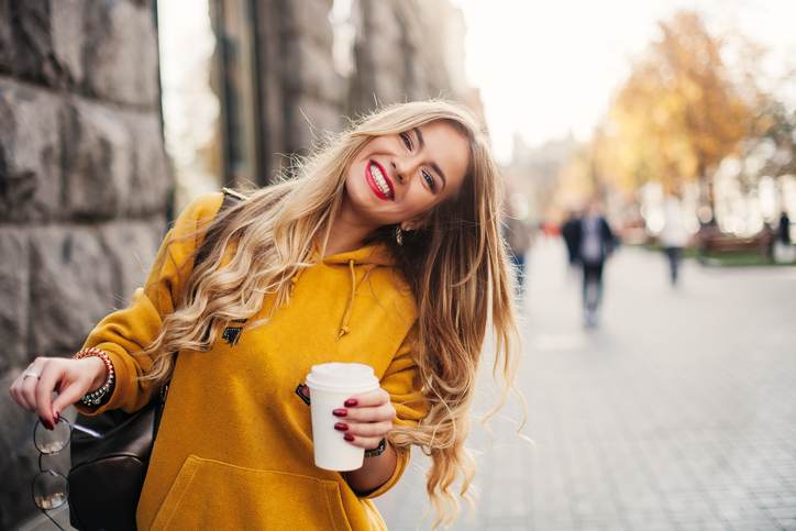 Stylish happy young woman wearing boyfrend jeans, white sneakers bright yellow sweetshot.She holds coffee to go. portrait of smiling girl in sunglasses and with bag